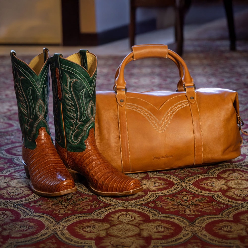 A pair of the Dinero 13" Caiman western boot in brown and green next to the Caori duffel bag in tan.